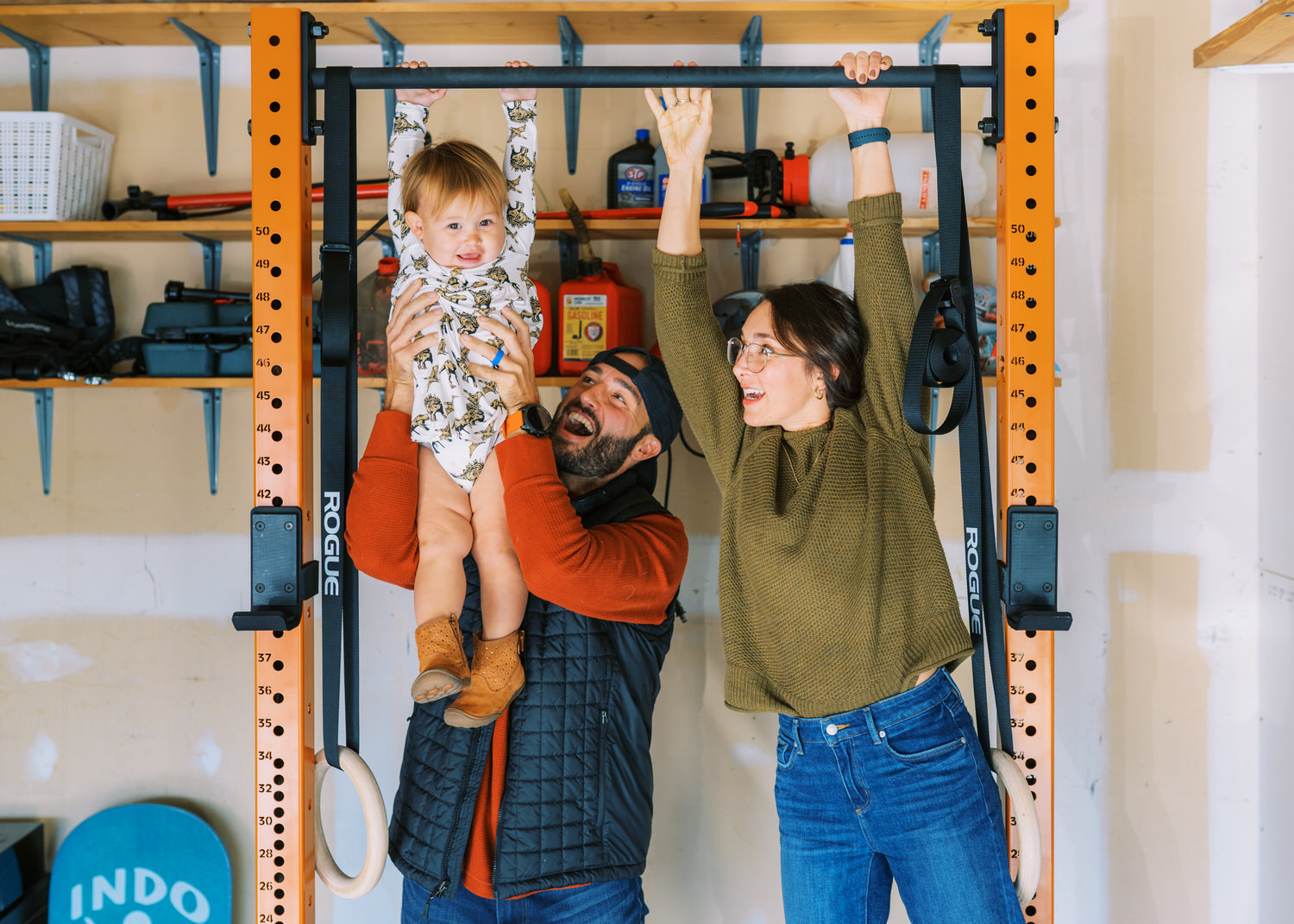 A toddler girl is lifted onto a pull-up bar by her dad, while her mom cheers her on