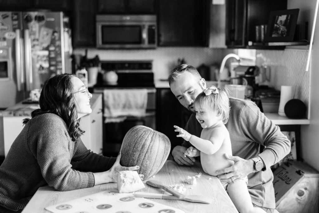 A toddler girl sticks her hand into a pumpkin at the kitchen table