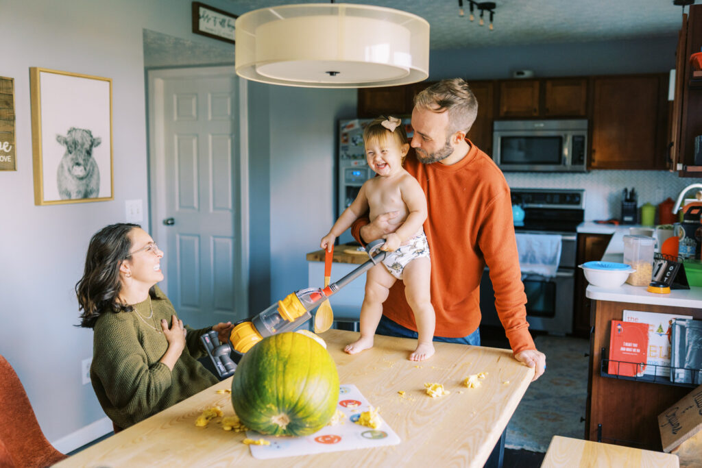 A toddler girl with a toy vacuum standing on a table while her parents laugh