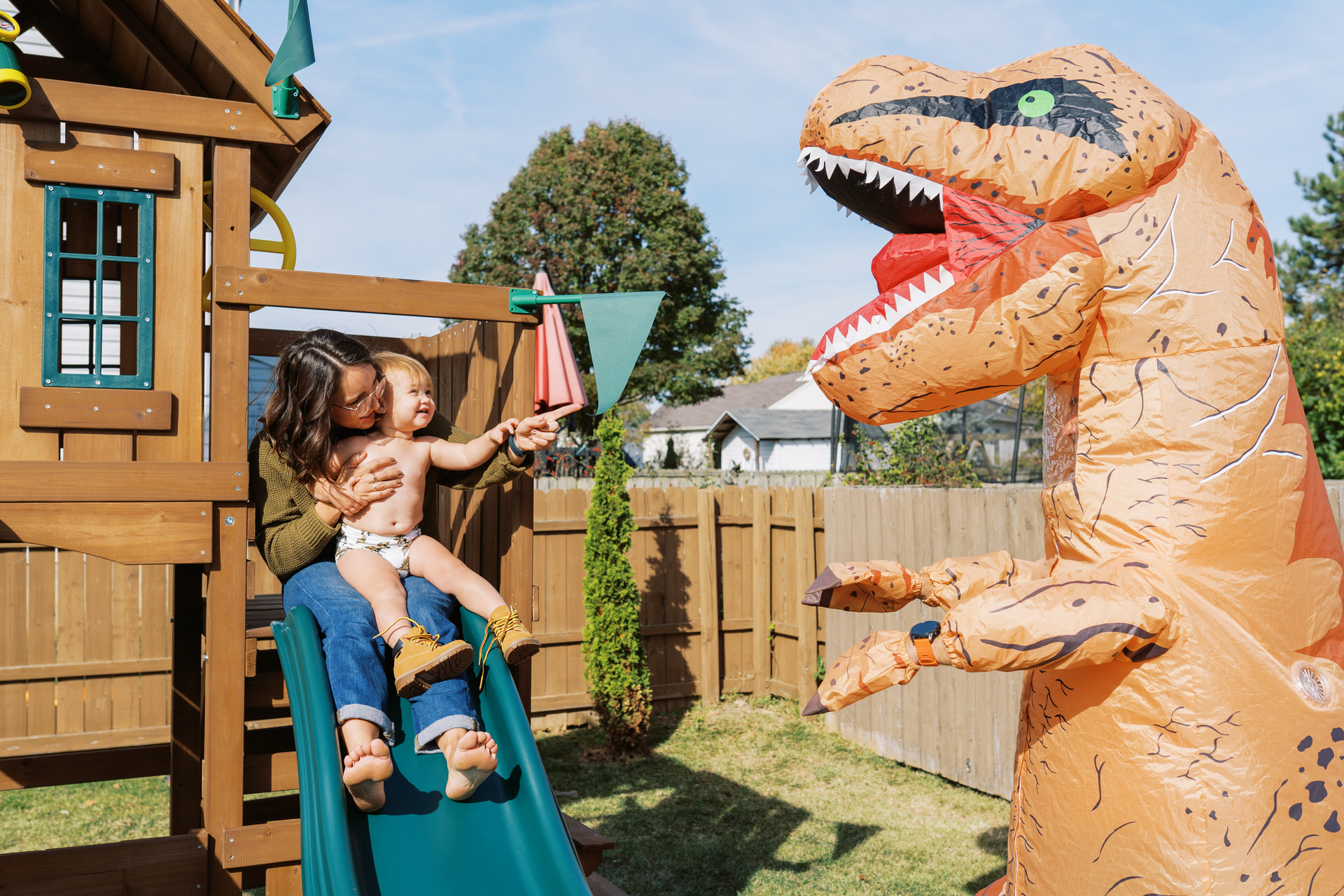 A mom and her toddler daughter sitting at the top of a slide while a person in a dinosaur costume approaches in a silly way