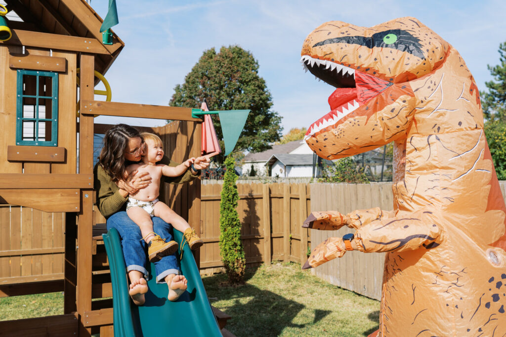 A family standing on top of a back yard playset together. The dad is wearing a dinosaur costume.