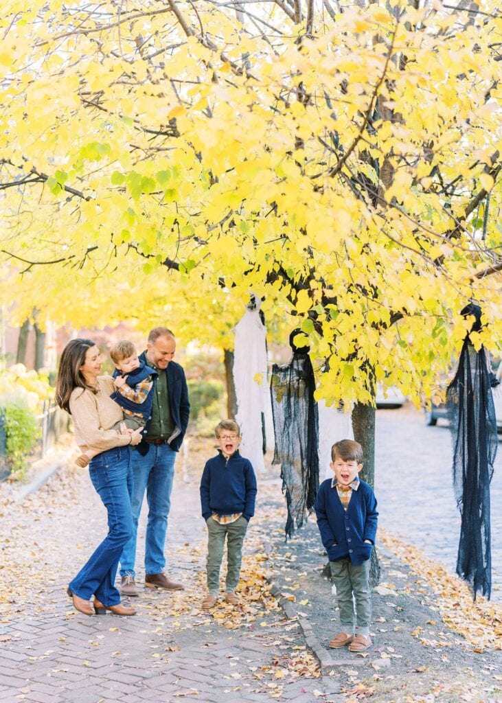 A family with 3 young boys standing on a sidewalk under a yellow tree 