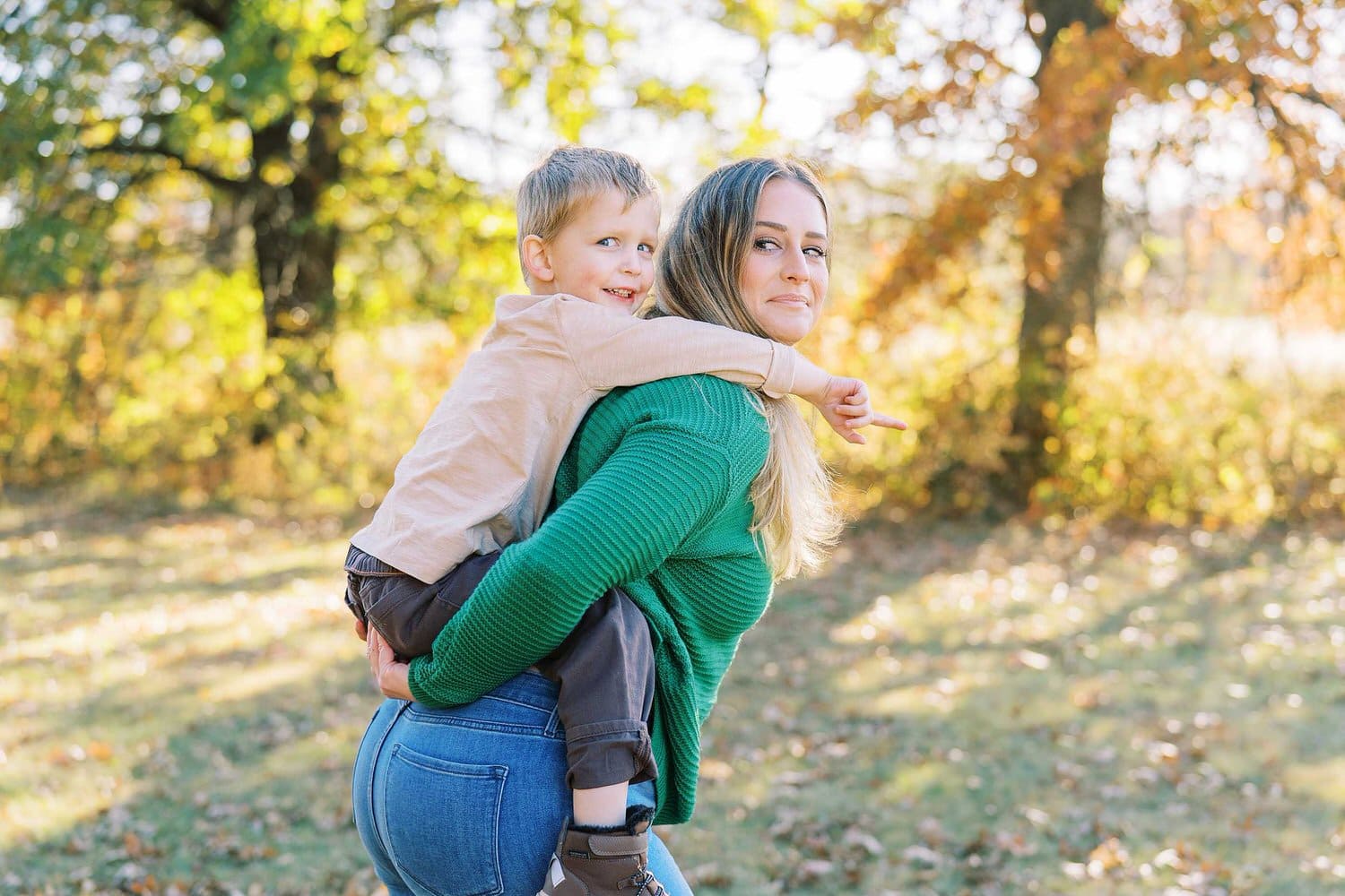 A mom in an emerald green sweater gives her toddler son a piggy back ride at a park in Columbus Ohio