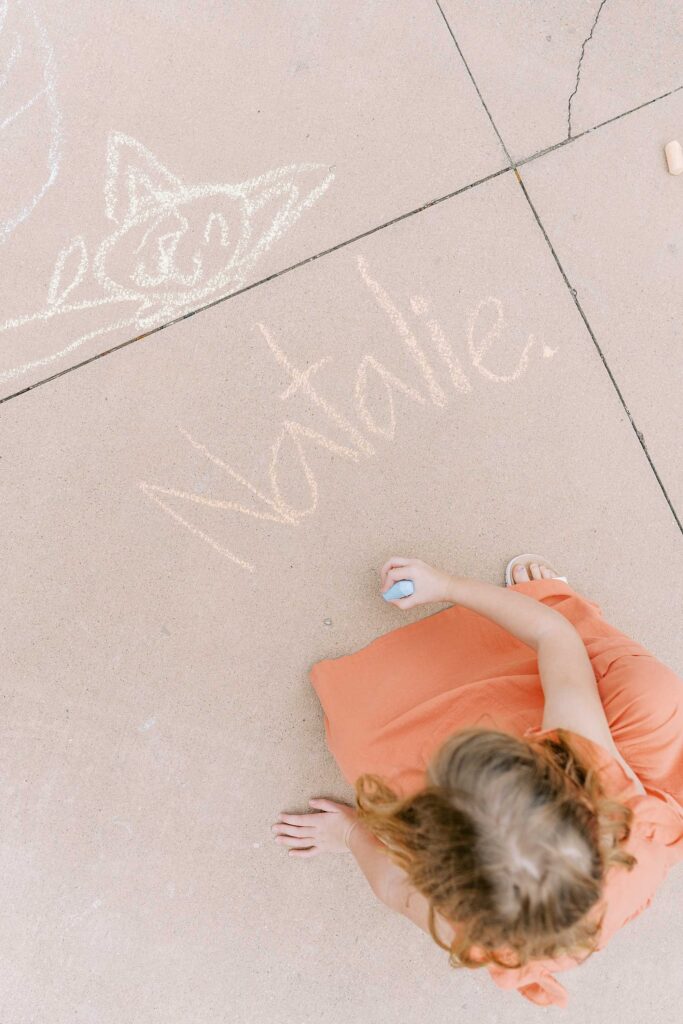 A little girl writes her name "Natalie" in sidewalk chalk