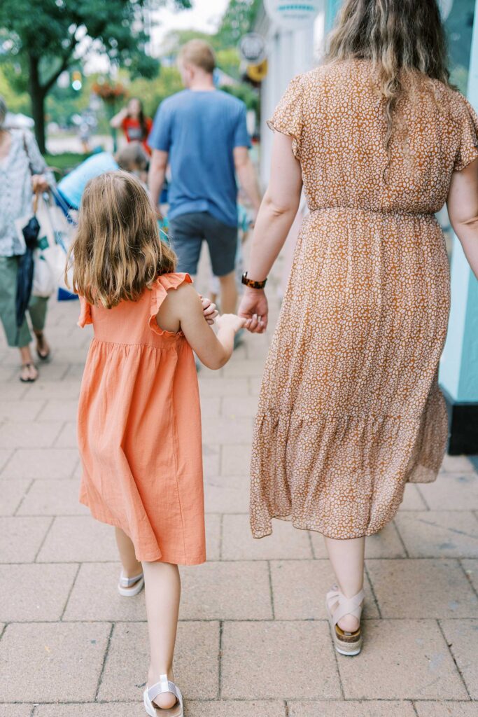 A little girl holds onto her mom's hand while they walk on a sidewalk