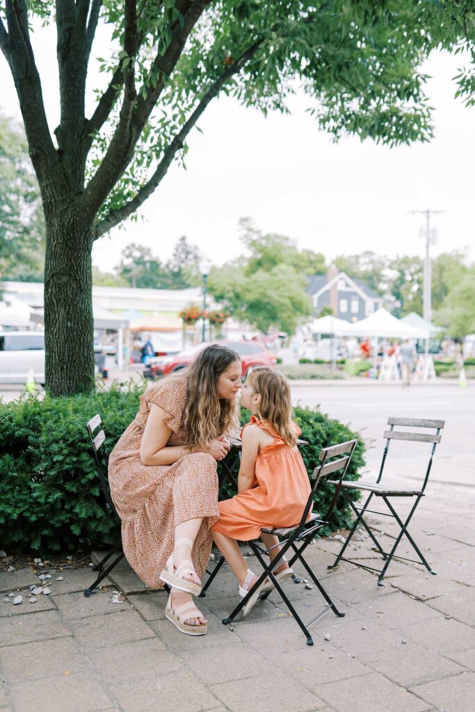 A mom and daughter touch noses as they sit at an outdoor cafe table