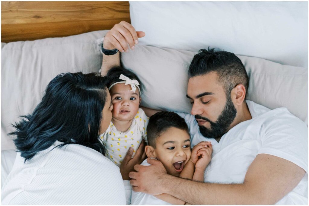 A family with a mom, a dad, a baby girl, and a young boy cuddles in their white pillows and bedsheets