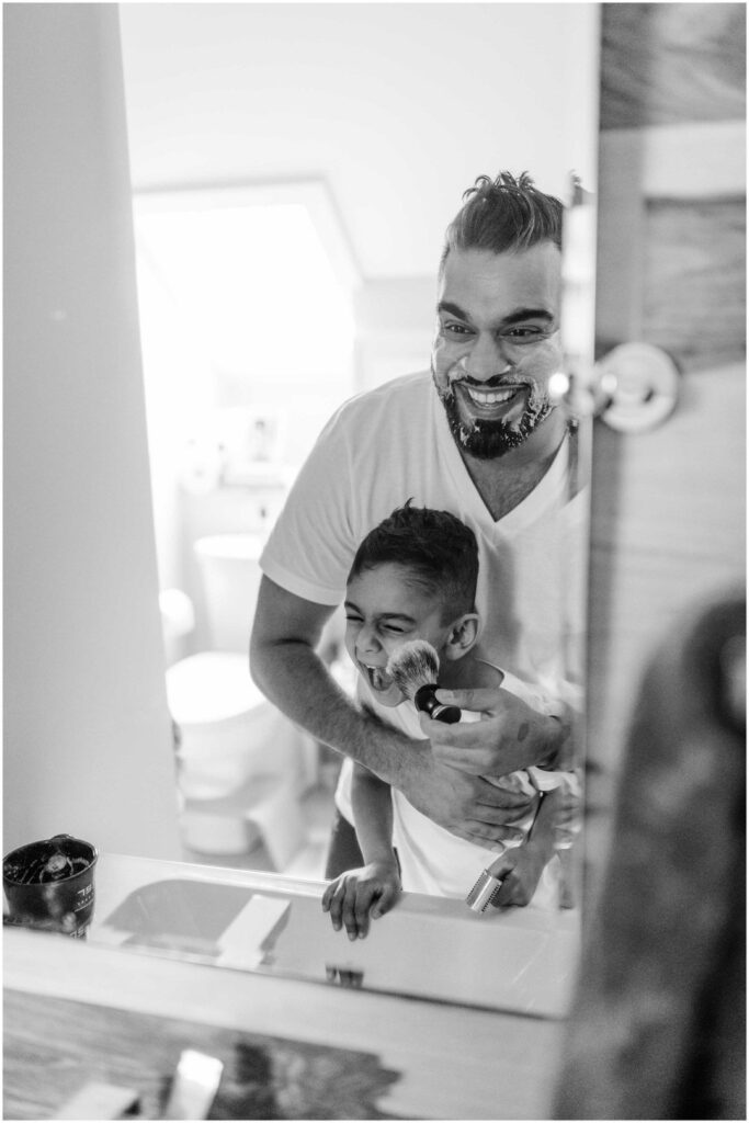 A dad and his young son playfully shave together while looking at their reflections in the mirror
