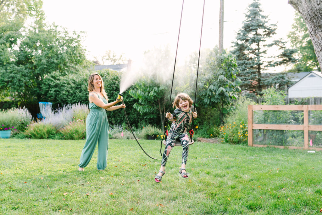 Sarah sprinkles Maddy with the hose in their backyard, captured by lifestyle photographer Daisy Zimmer