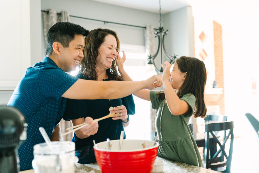a mom and dad playfully rub flour on their daughter's nose while baking together in the kitchen