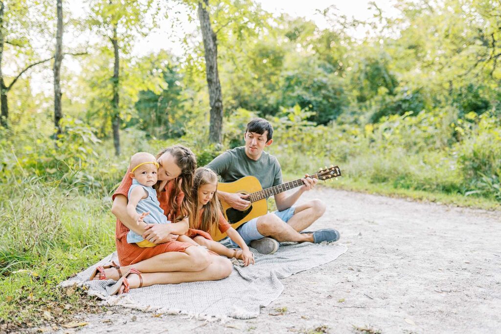 Parents and their two young daughters sitting on the ground while the dad plays a guitar