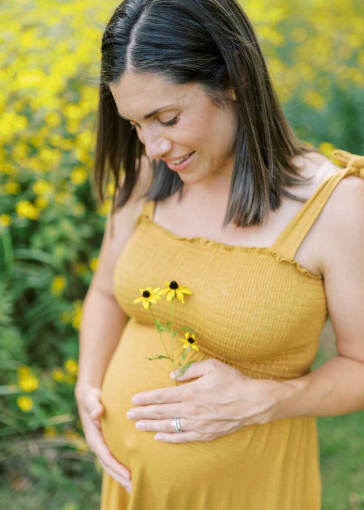 A pregnant lady with dark hair and wearing a yellow dress holding a black eyed susan flower and looking down at her belly
