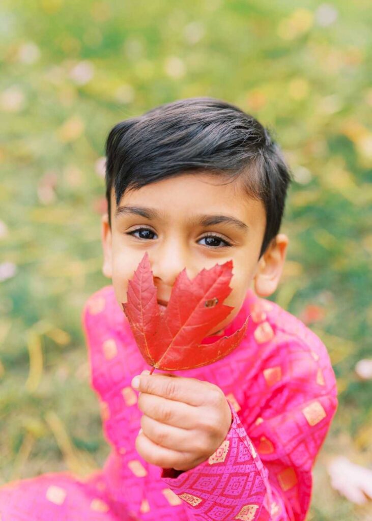 A young Indian boy with dark eyes and long eyelashes covering the bottom half of his face with a red leaf 