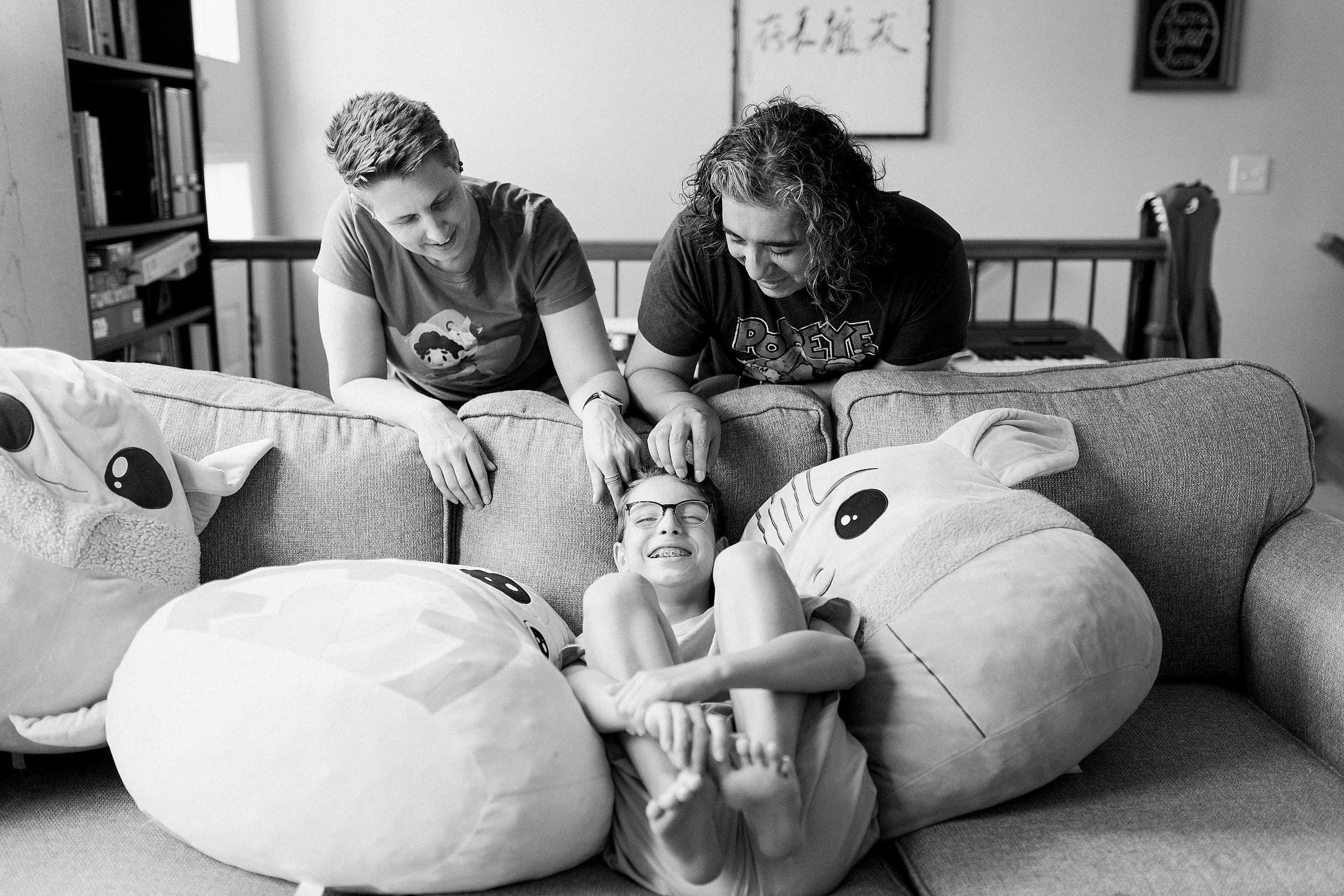 Jack sits between two huge squishmallow pillows on the couch while his parents play with his hair