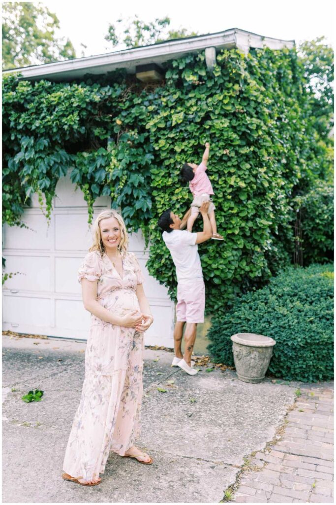 Dad lifts his son to pick a leaf from an ivy wall. Mom stands in front of them holding her pregnant belly