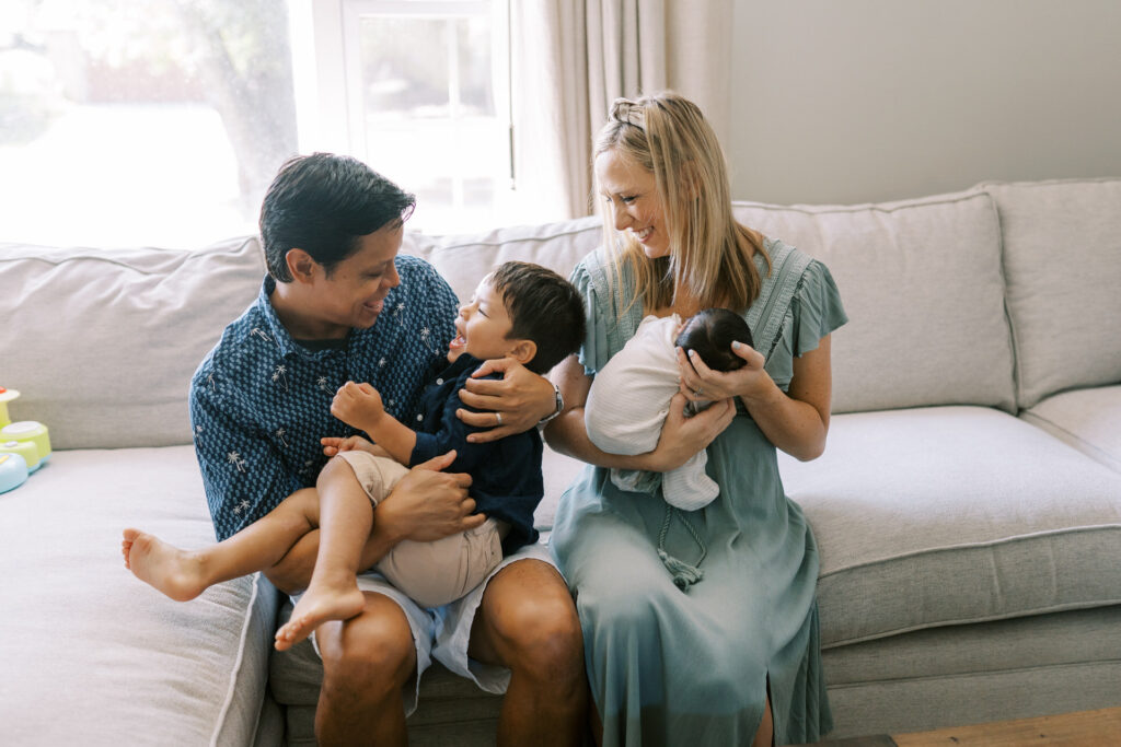A family with a toddler son and a newborn baby sit together laughing on the couch