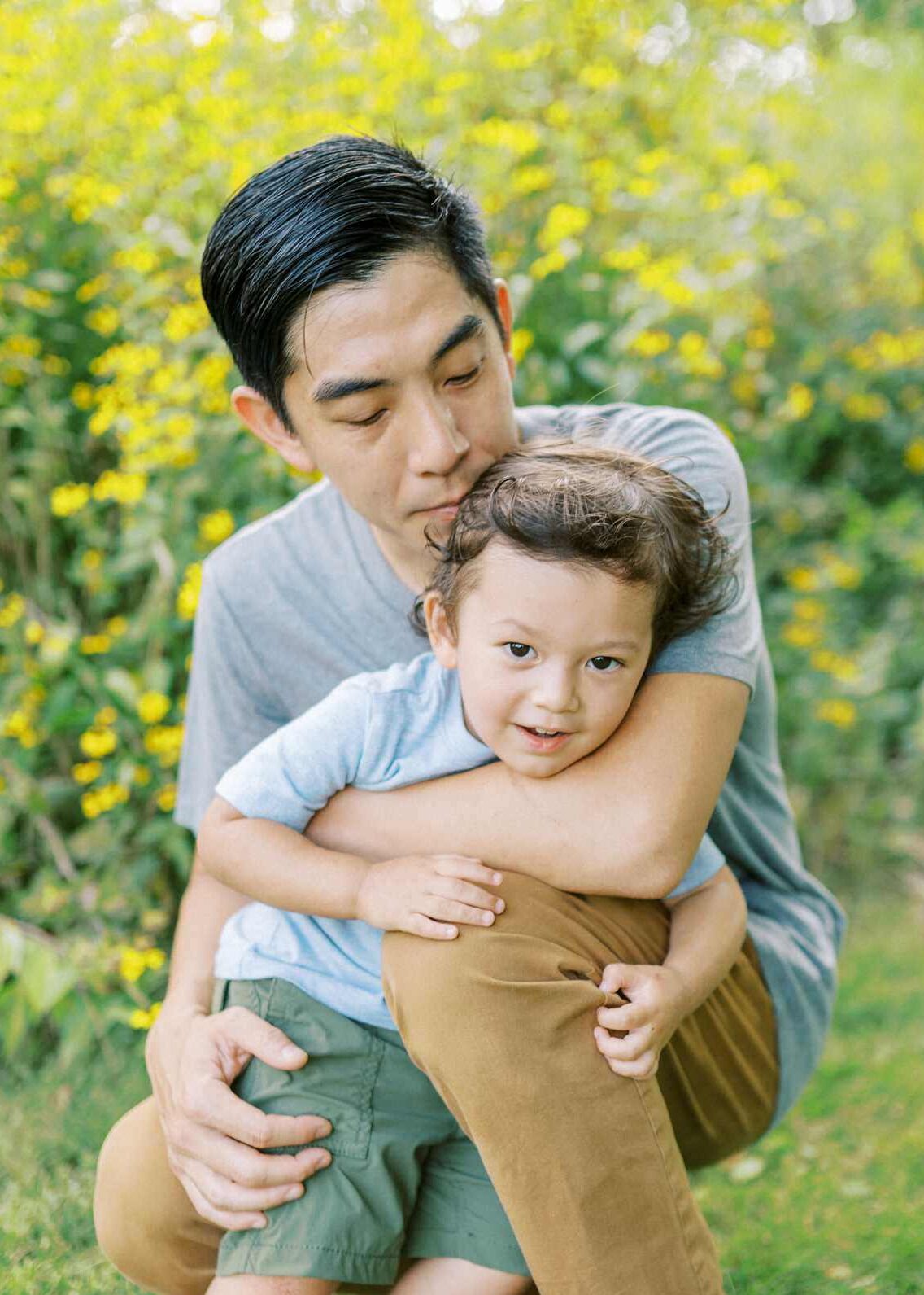 a dad hugs his toddler son in a field of yellow flowers