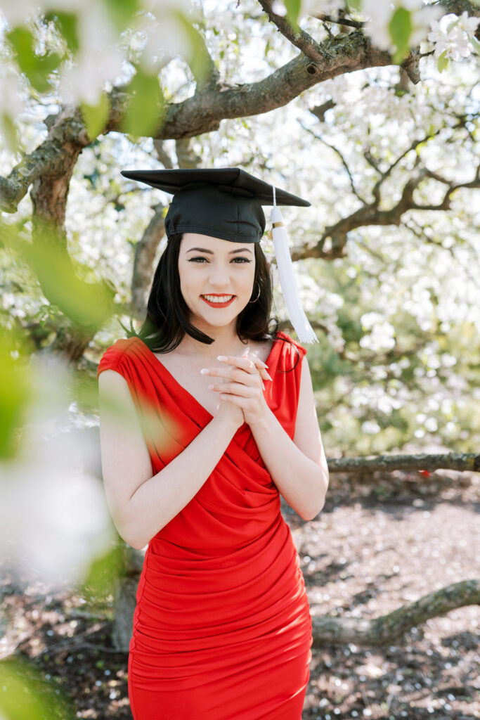 A college girl wearing a red dress and her graduation cap in front of a flowering tree