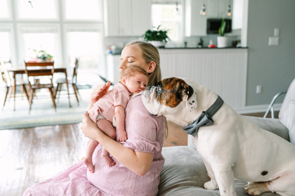 Maggie holds her newborn daughter while a bulldog wearing a bowtie sniffs the baby's face