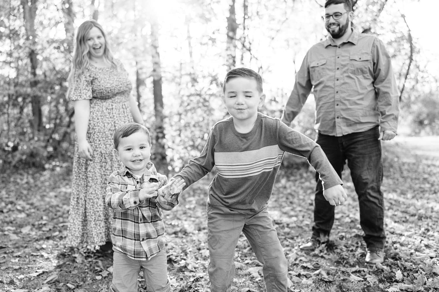 Two young boys having a dance party while their mom and dad laugh, A family with two young sons sits on a blanket in front of autumn trees, captured during a fall mini session by Daisy Zimmer Photography