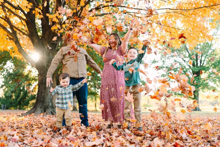 A family with two young boys tosses colorful autumn leaves in the air, A family with two young sons sits on a blanket in front of autumn trees, captured during a fall mini session by Daisy Zimmer Photography