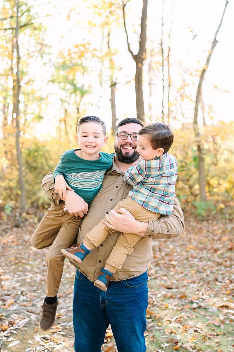 A dad playfully picks up his two young sons in front of colorful autumn trees