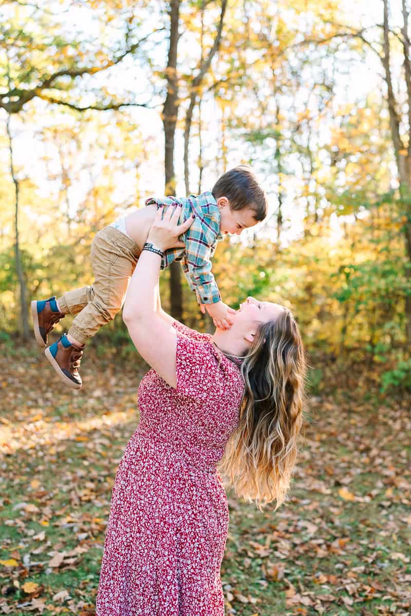 A mom with long brown hair lifts her young son into the air