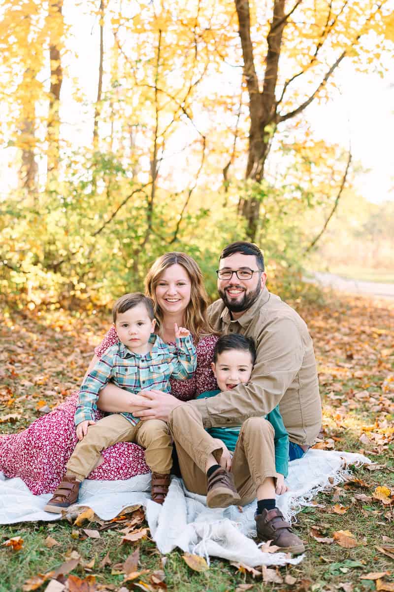 A family with two young sons sits on a blanket in front of autumn trees, captured during a fall mini session by Daisy Zimmer Photography