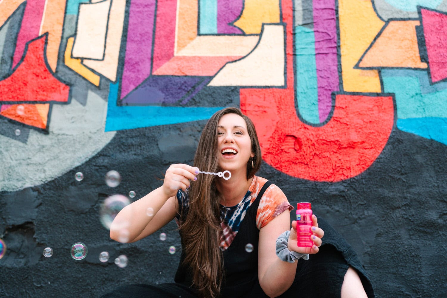 Daisy, a Columbus Ohio Family Photographer, is wearing a tie-dye shirt and blowing bubbles in front of a colorful painted wall 